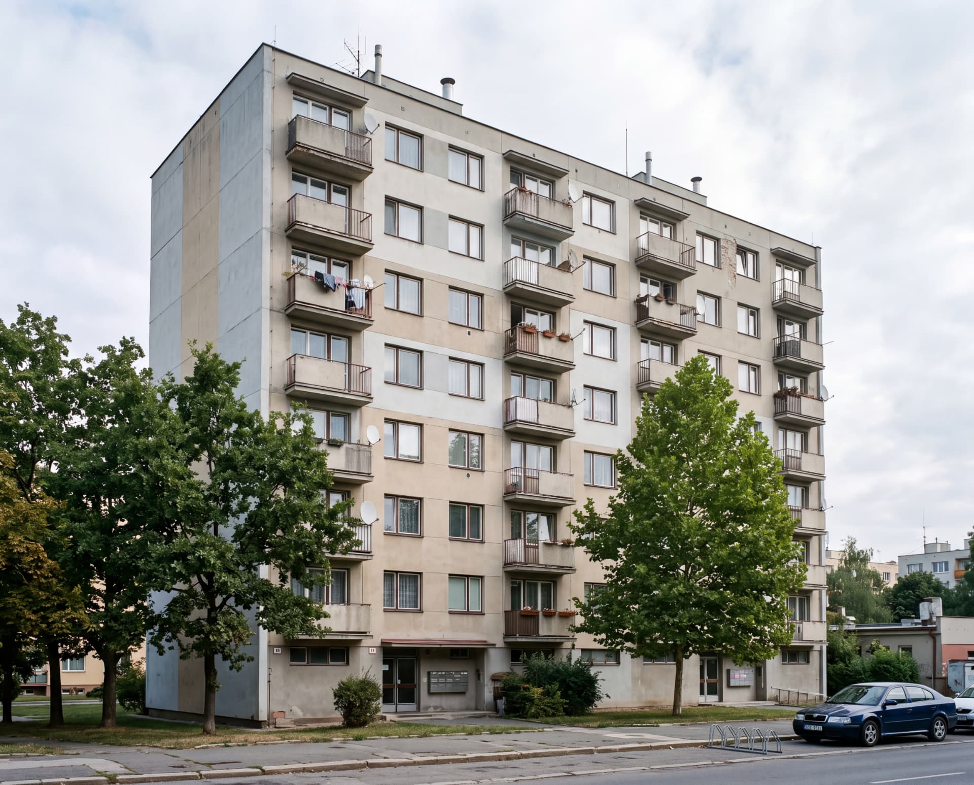 Osmipodlažní panelový dům s balkony, satelitními anténami a zelení kolem vchodů — typická nemovitost vhodná k výkupu ve stavu, v jakém je
